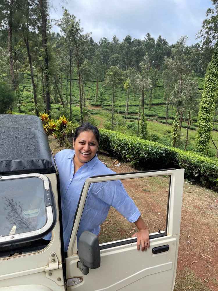 Namita, owner of Mysore Plantations, smiling beside a jeep with tea and pepper hills behind her